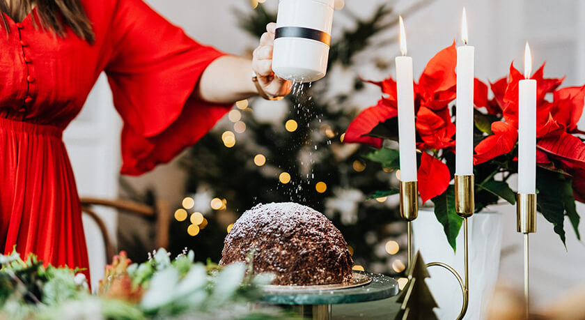 woman in red shaking icing sugar over christmas pudding on table with candles and red poinsettia