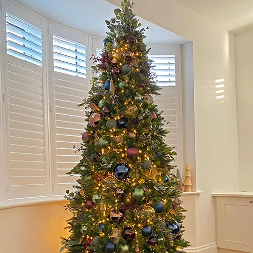 jewel tone decorated slim green christmas tree in white lounge with white roman blinds
