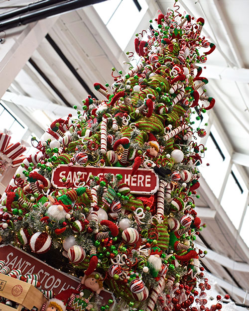 large tree filled with elf christmas decorations in red, white, and green with santa stop here sign