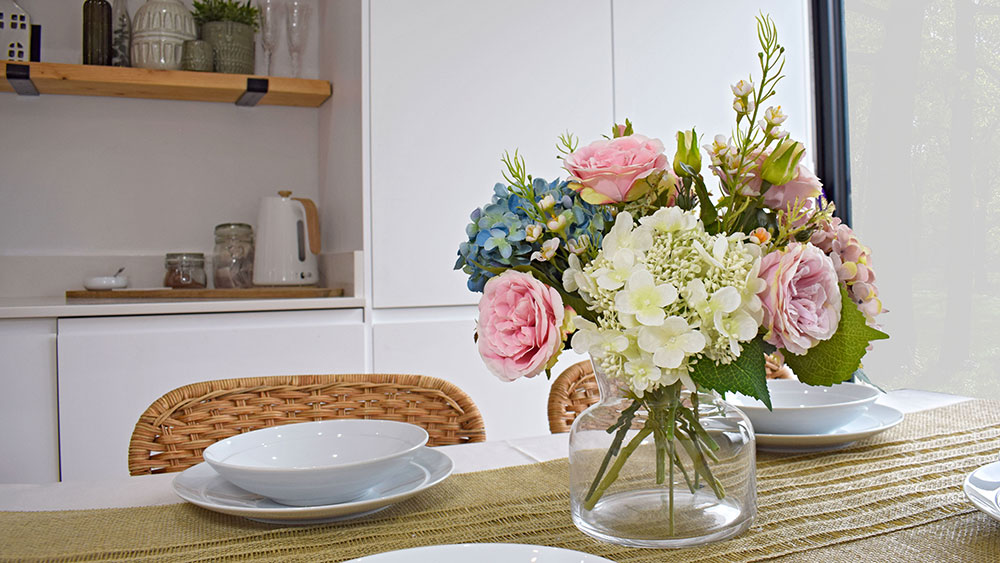 pastel flower trend - collection of pastel pink roses, blue hydrangeas, and cream flowers in glass vase on dining table