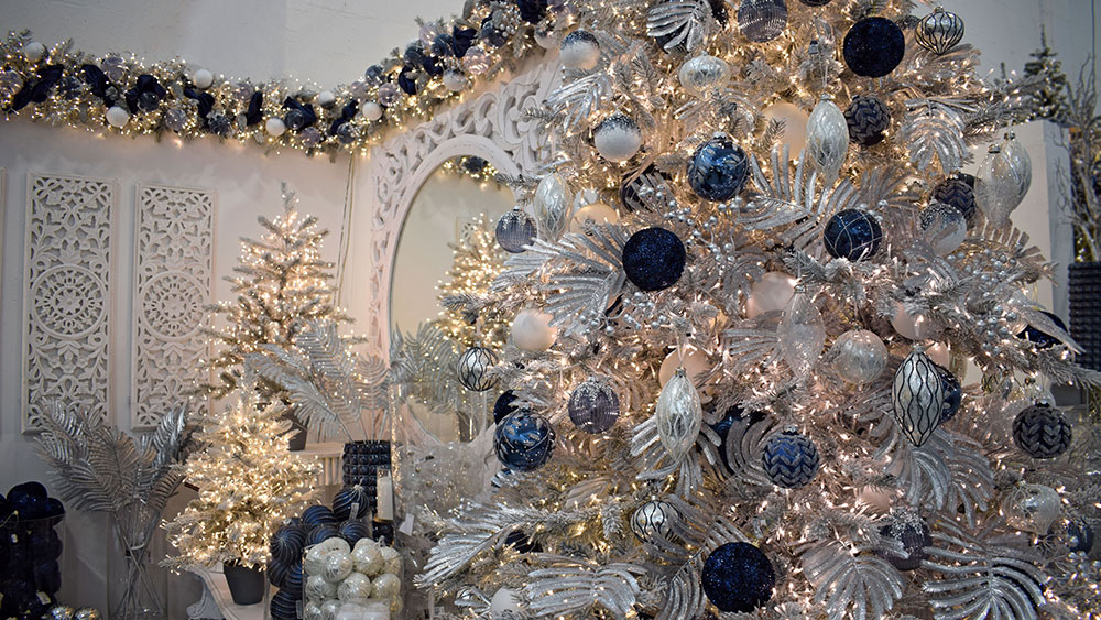 white christmas tree with midnight blue christmas decorations, silver faux ferns and clear baubles with festive display in background
