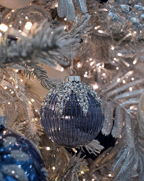 close up of dark blue ribbed bauble with silver sequine and glitter on top hanging from the white christmas tree