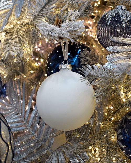close up of mat white bauble with midnight blue christmas decorations hanging in tree with lights