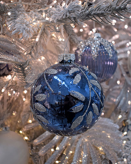 close up of midnight blue bauble with glitter leaf design hanging in white tree