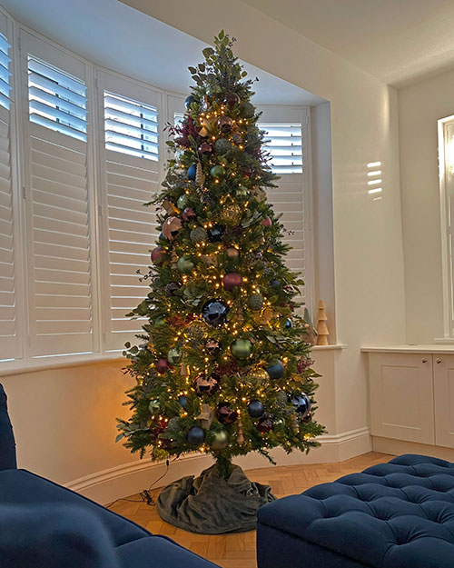 slim green christmas tree with purple, dark blue, green, wood, and gold decorations next to a dark blue sofa and ottoman