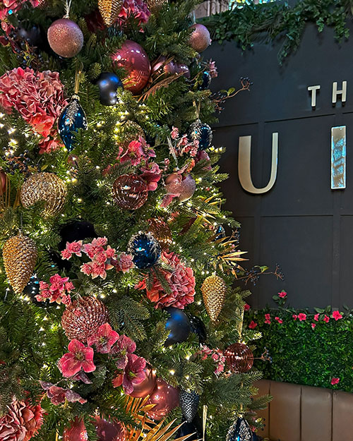 close up of pink faux hydrangeas, gold pine cones, blue baubles on a large green christmas tree next to the venue sign
