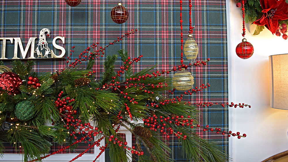 traditional christmas decorations - red berries and green foliage garland over fireplace with tartan wallpaper and baubles