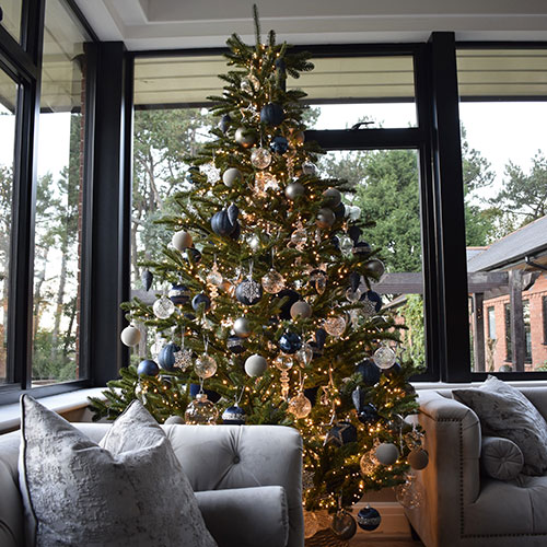 christmas tree with blue and clear decorations next to windows and grey sofas